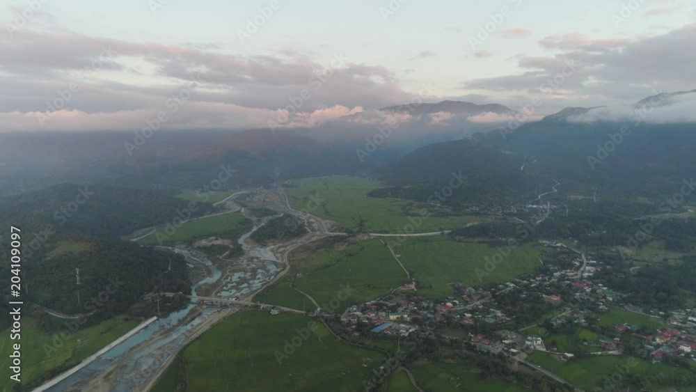 Aerial view of farmer fields, river, village in the background of mountains at sunset on the island of Luzon, Philippines. Mountain landscape with valley and village. Aerial view of farmer fields, river, village in the background of mountains at sunset on the island of Luzon, Philippines. Mountain landscape with valley and village.