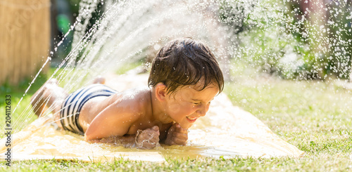 Boy cooling down with garden hose, family in the background on a hot summer day photo