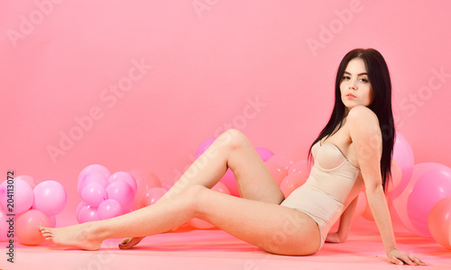 Girl on pensive face looks attractive. Fashion model in slinky clothes with slim figure, young and healthy. Woman in beige bustier, bodysuit posing near air balloons, pink background. Fashion concept.