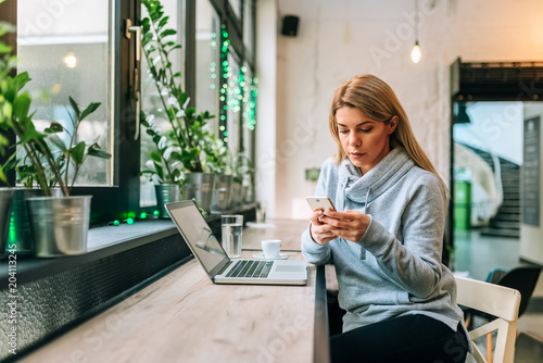 Serious blonde woman in casual clothing using phone and laptop at the cafe. photo