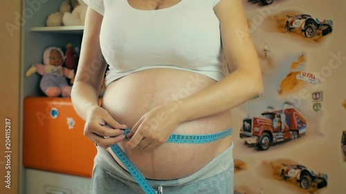 Young pregnant girl measures the volume of her belly in the children room in slow motion