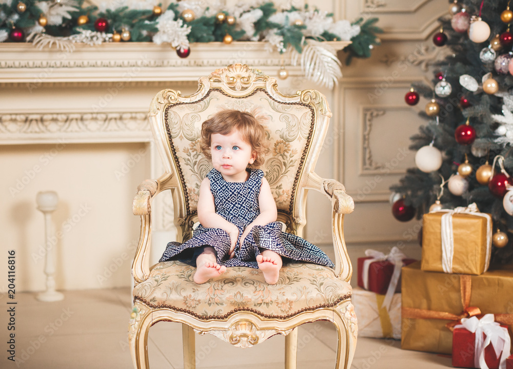 Cute small girl in beautiful dress in chair.Christmas tree background.Studio shooting.Holiday concept.