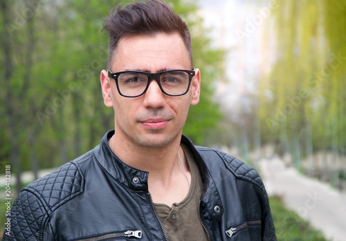 Handsome man of 25 years. A young man in glasses and a leather jacket in the background of a park in the city.