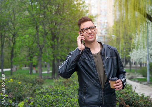 Man is talking on a smartphone. Drink coffee on the street and talk on the phone. A modern stylish man with glasses and a leather jacket.