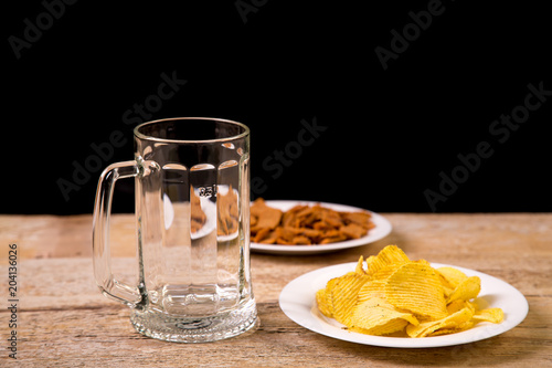 On a wooden table is a mug for beer. Nearby is a white plate with potato chips. Horizontal photo.
