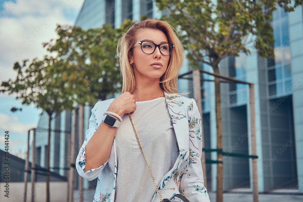 Beautiful fashionable woman in stylish clothes and glasses with a handbag, standing against a skyscraper.