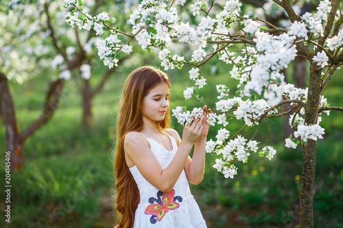 A portrait of a beautiful young girl with blue eyes in a white dress in the garden with apple trees blosoming having fun and enjoying smell of flowering spring garden at the sunset