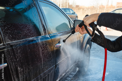 Girl washes a black SUV with a high-pressure car wash. Soap foam and water on the machine. Self-service car wash. A young woman washes the car in the street car wash in the fresh air.