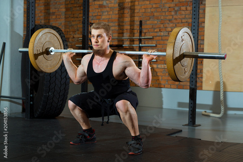 a handsome young man with a beard is engaged in squatting with a barbell in the hall of a cross