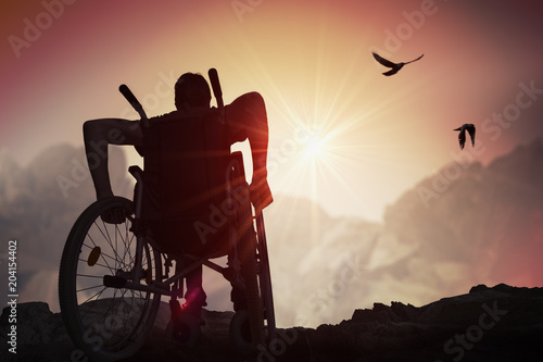 Disabled handicapped man has a hope. He is sitting on wheelchair and stretching hands at sunset.