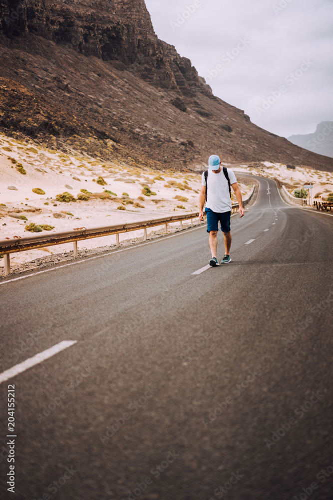 Traveler with backpack standing in the center of an epic winding road. Huge volcanic mountains in the distance behind him. Sao Vicente Cape Verde Traveler with backpack standing in the center of an epic winding road. Huge volcanic mountains in the distance behind him. Sao Vicente Cape Verde