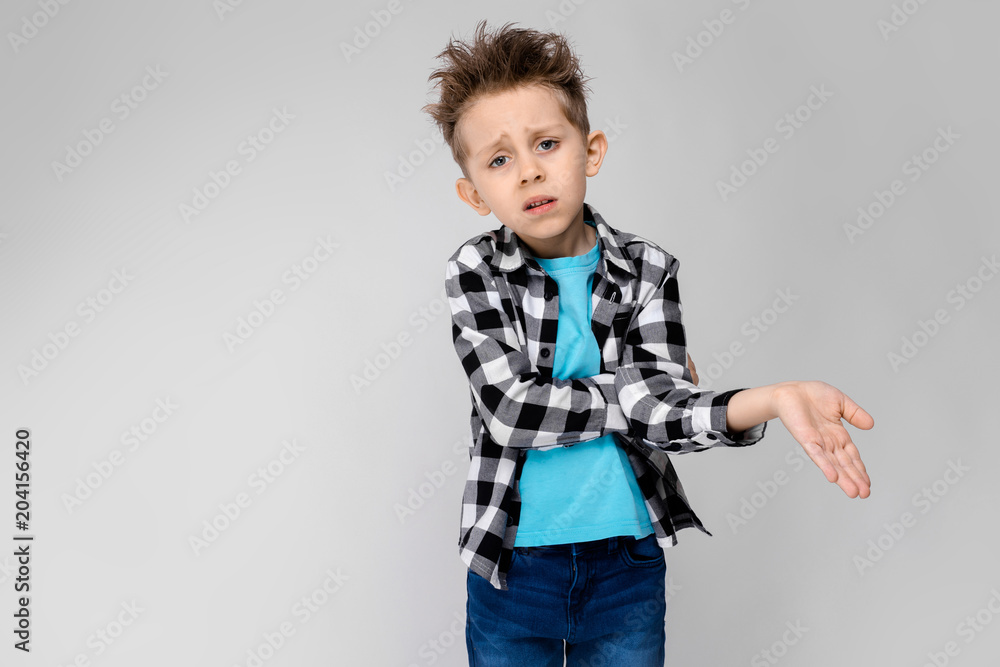 A handsome boy in a plaid shirt, blue shirt and jeans stands on a gray background. The boy folded his arms over his chest. The boy is unhappy. The boy shows his hand A handsome boy in a plaid shirt, blue shirt and jeans stands on a gray background. The boy folded his arms over his chest. The boy is unhappy. The boy shows his hand