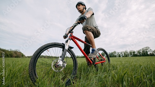 Bicycle sports, traveling, healthy lifestyle and activity. Low angle view of young man riding bicycle on a meadow with high green grass