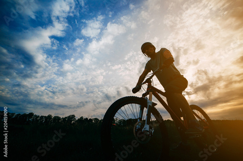 Sports, tourism and activity concept. Silhouette of a cyclist in helmet riding a bicycle over sunset sky background