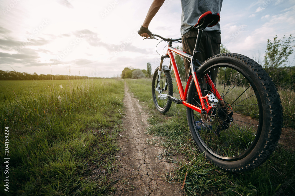 Low angle view of young man in helmet standing with bicycle at country road enjoying sunset. Tourism, healthy lifestyle and activity