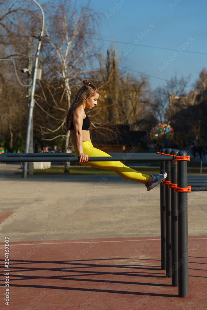 Girl on street workout. She does gymnastic exercises on parallel crossbars. Girl trains strength and coordination. She is dressed in black topic and yellow sports pants.