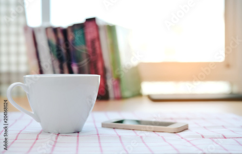 A cup of coffee and a smartphone is on the tablecloth and the book is blurred as the background.