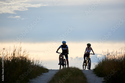 Couple of cyclists sitting on bicycles on a country road watching each other in the background a horizon with a beautiful evening sky. The guys are dressed in sports clothes and helmets