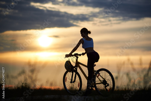 Back view of slender female cyclists sitting on bicycle and posing in sportswear. Sporty woman riding bike, enjoying and observing amazing and fantastic view of sunset and landscape.