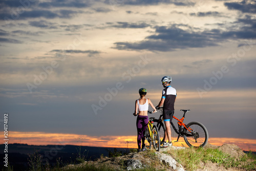 Couple standing together near their mountain bikes on rock hill and observing beautiful view of landscapes, sunset and cloudy sky. Athletic cyclists wearing helmets and sportswear.