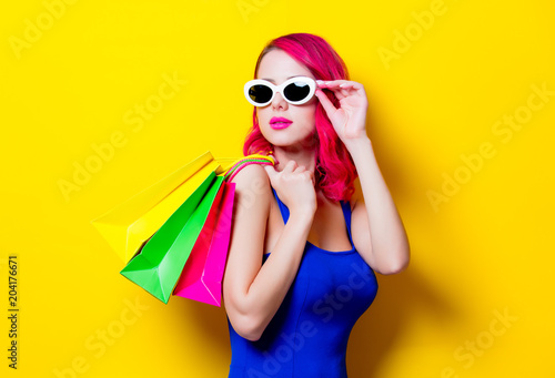Young pink hair girl in blue dress with colored shopping bags. Portrait isolated on yellow background