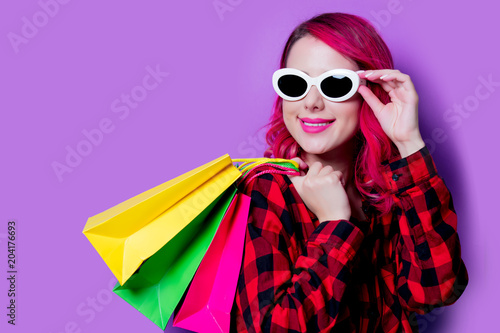 Young pink hair girl in red tartan shirt with shopping bags and sunglasses. Portrait isolated on purple background