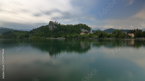 Panoramiczny widok Bled, Słowenia, jezioro, skaliste wzgórze z ruinami  średniowiecznego zamku, poniżej wieże kościoła, zieleń i niebo odbija się w spokojnej tafli wody © Wioletta