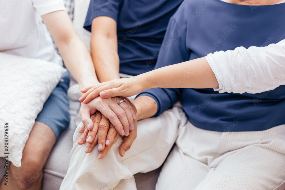 Obraz premium Close up of family with adult children and senior parents putting hands together and sitting on sofa at home together. Family unity and cooperation concept