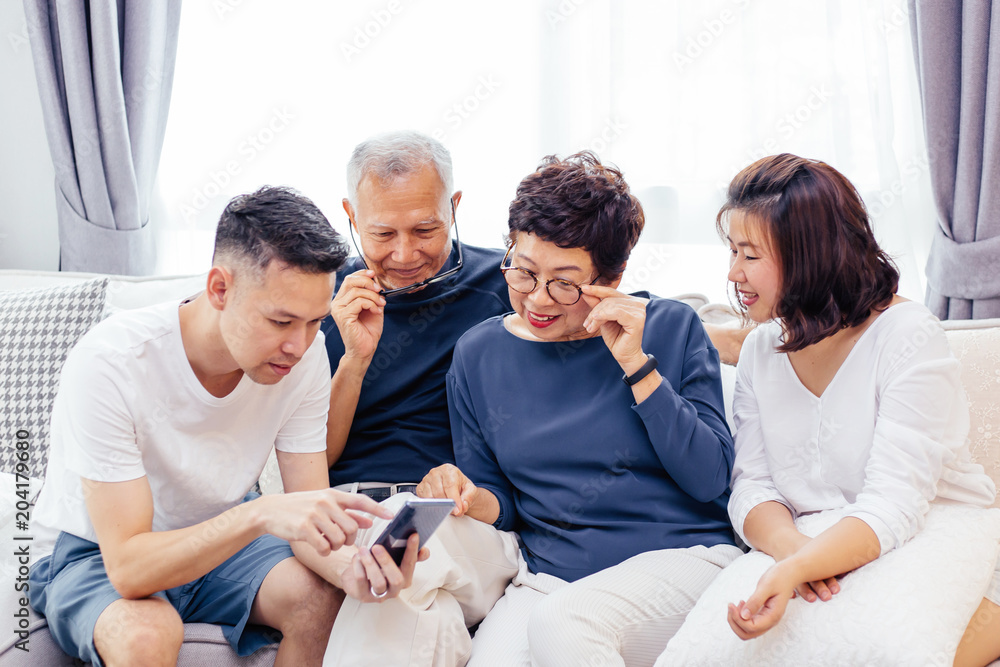 Fototapeta premium Asian family with adult children and senior parents using a mobile phone and relaxing on a sofa at home together