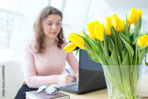 Bouquet of yellow tulip flowers is on the desktop. Congratulation of the employee on his birthday. Young woman is studying while sitting at a table. Woman working at office. #204199424