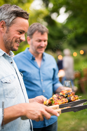 in a summer evening   two men  in their forties prepares a barbecue for  friends gathered around a table in the garden