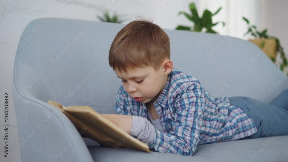 Cute little boy in casual clothes is reading funny book aloud lying on comfortable sofa and laughing. Happy childhood, preschool education and hobby concept. Cute little boy in casual clothes is reading funny book aloud lying on comfortable sofa and laughing. Happy childhood, preschool education and hobby concept.