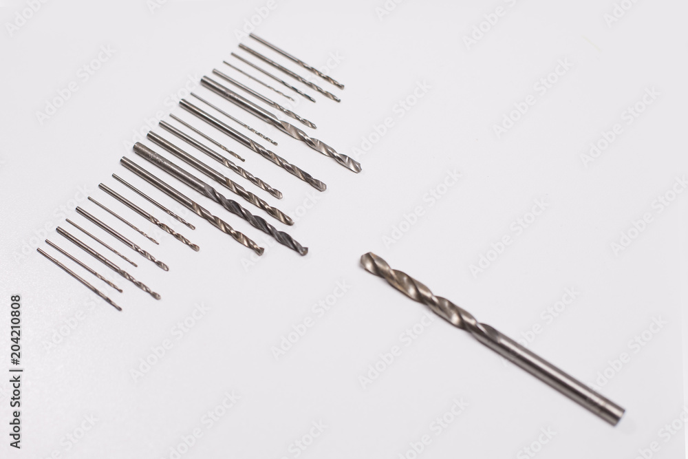 Large and small drills for drilling lie in a row on a white isolated background. Opposite them a large drill bit. View from above. Close photo Large and small drills for drilling lie in a row on a white isolated background. Opposite them a large drill bit. View from above. Close photo