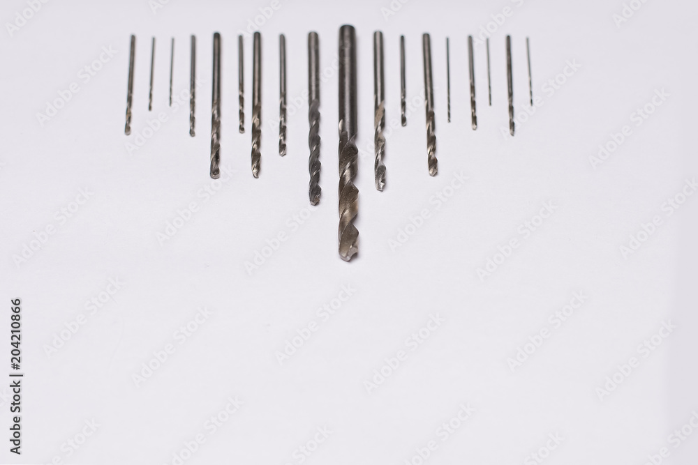 Large and small drills for drilling lie in a row on a white isolated background. Shallow depth of field. Focus on the tip of a large drill. View from above. Large and small drills for drilling lie in a row on a white isolated background. Shallow depth of field. Focus on the tip of a large drill. View from above.