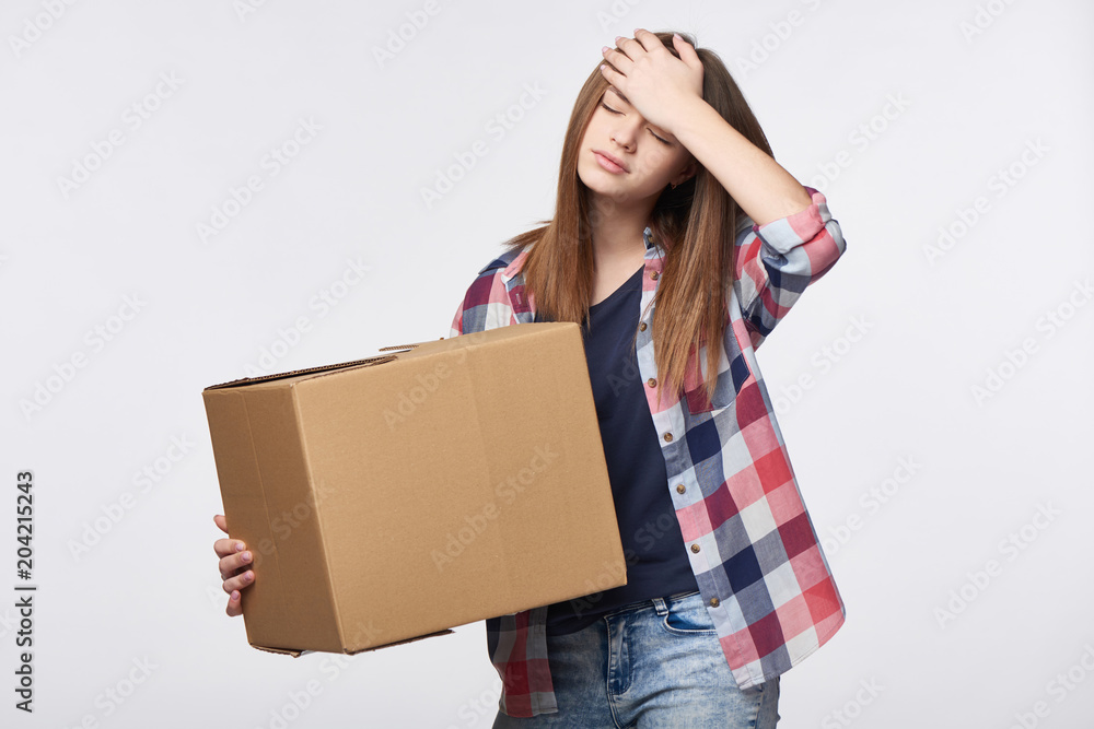 Delivery, relocation and unpacking problems. Discontent young woman holding cardboard box with hand on head Delivery, relocation and unpacking problems. Discontent young woman holding cardboard box with hand on head