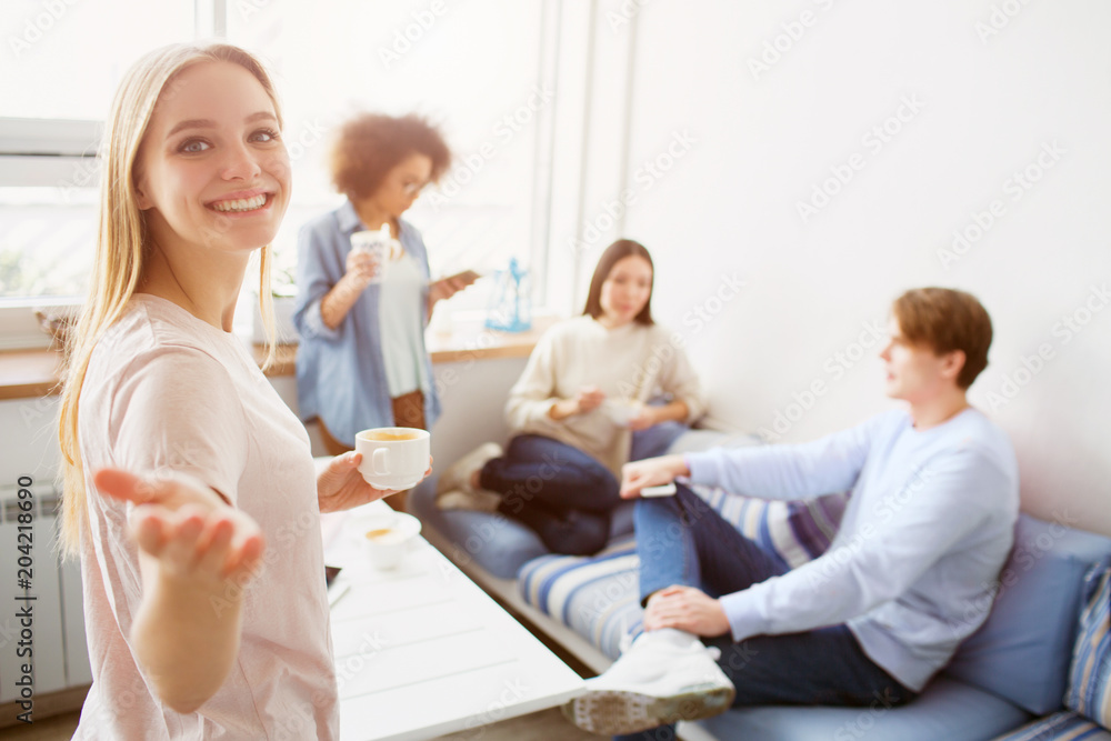 Nice picture of blonde that is looking to camera and smiling. She is pointing on it and holding a cup of coffee while her friends are talking with each other and drinking coffee.