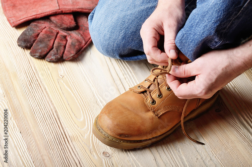 Carpenter in blue jeans tying shoelaces of yellow work boots on on wooden floor. Place for text
