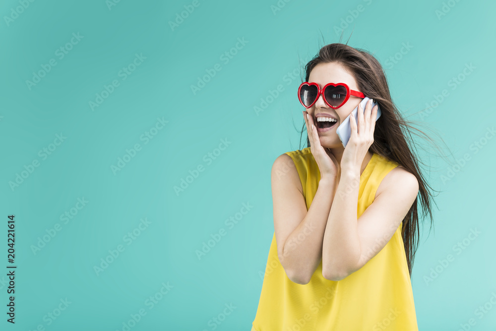 Fototapeta premium Beautiful smiling brunette girl wears yellow t-shirt and sunglasses talking by phone before blue background