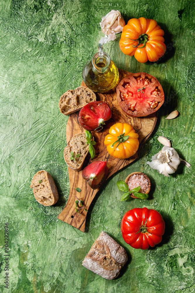 Variety of red and yellow organic tomatoes with olive oil, garlic, salt and bread for salad or bruschetta on wooden cutting board over green texture background. Top view, space.