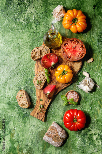 Variety of red and yellow organic tomatoes with olive oil, garlic, salt and bread for salad or bruschetta on wooden cutting board over green texture background. Top view, space.