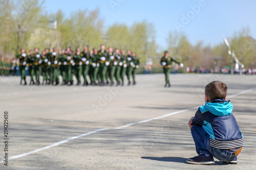 The boy sits on the ground and watches the military parade. A future soldier boy in jeans and a jacket looks at soldiers in the army marching on a military holiday.