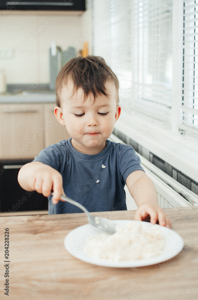 The little boy in the kitchen eagerly eating rice with a spoon independently The little boy in the kitchen eagerly eating rice with a spoon independently