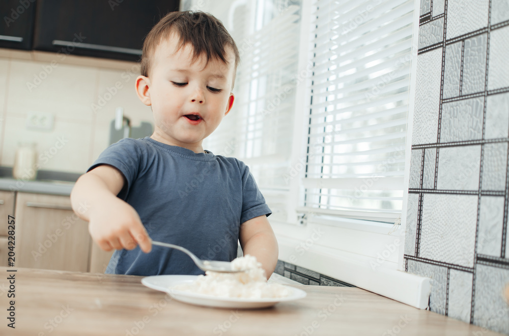 The little boy in the kitchen eagerly eating rice with a spoon independently The little boy in the kitchen eagerly eating rice with a spoon independently