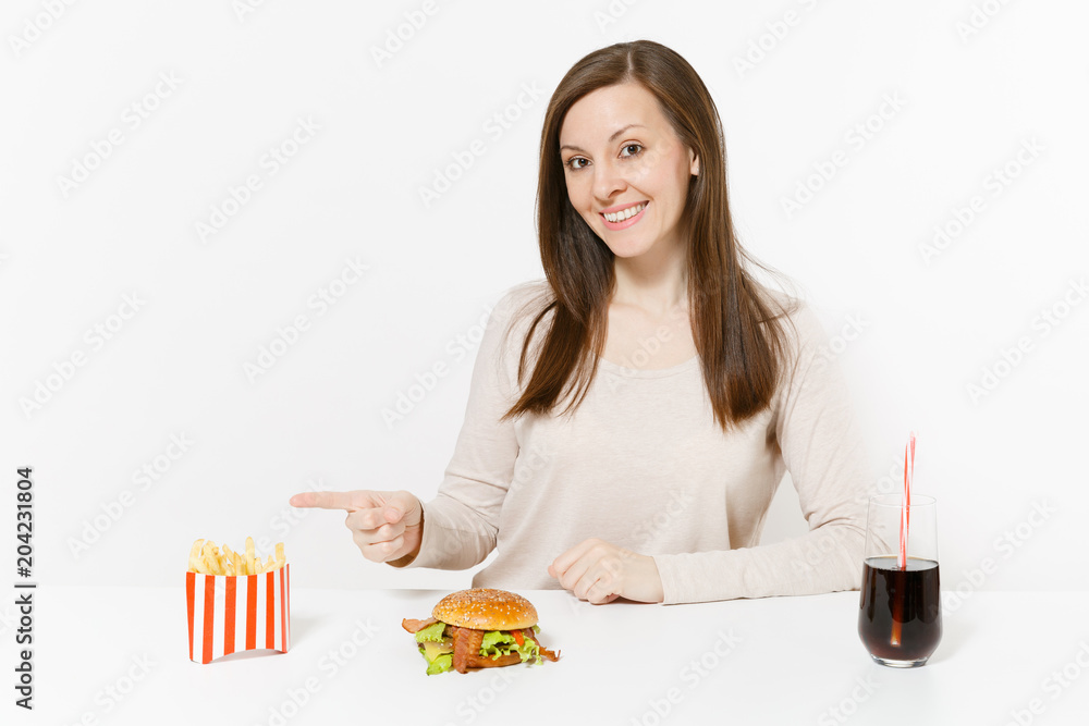 Beautiful young woman sitting at table with burger, french fries, cola in glass bottle isolated on white background. Proper nutrition or American classic fast food. Advertising area with copy space. Beautiful young woman sitting at table with burger, french fries, cola in glass bottle isolated on white background. Proper nutrition or American classic fast food. Advertising area with copy space.