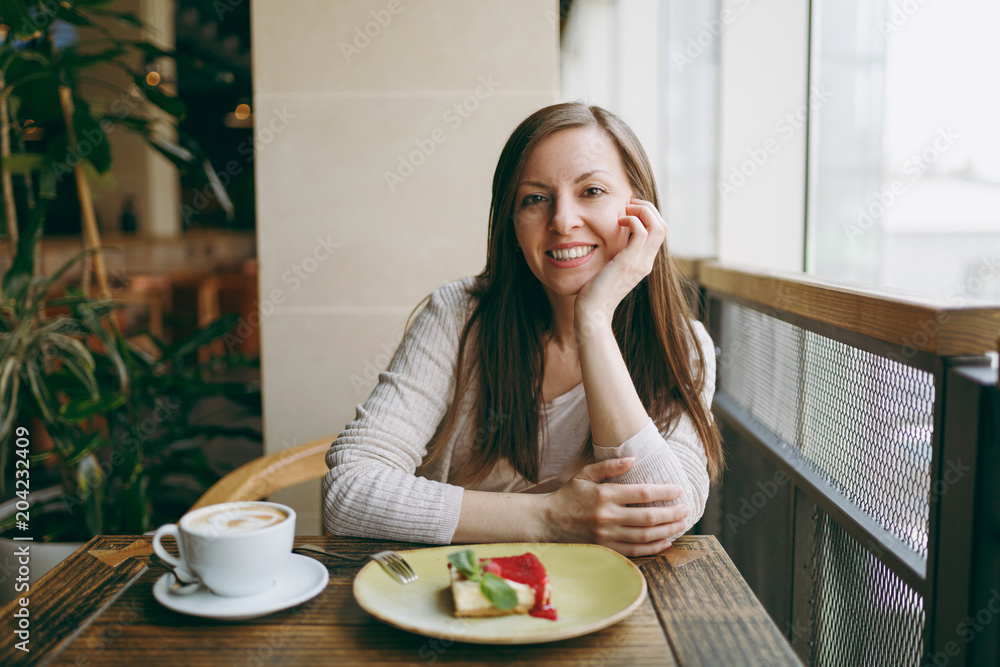 Attractive young woman sitting alone near big window in coffee shop at table with cup of cappuccino, cake, relaxing in restaurant during free time. Young female having rest in cafe. Lifestyle concept. Attractive young woman sitting alone near big window in coffee shop at table with cup of cappuccino, cake, relaxing in restaurant during free time. Young female having rest in cafe. Lifestyle concept.