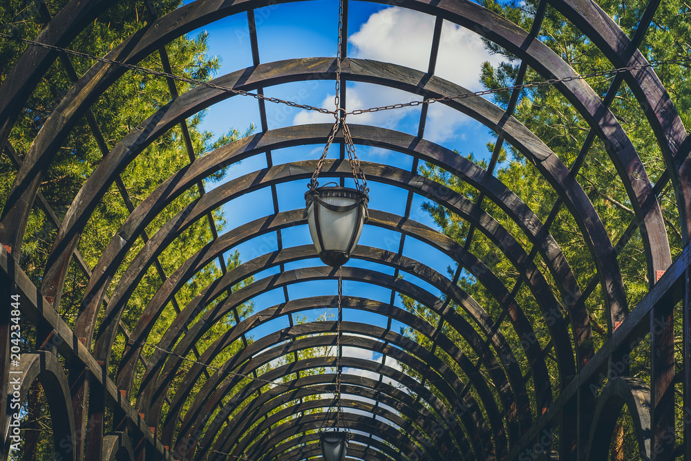 arch from a wooden brown beam with lanterns against a background of green trees and a blue sky with clouds arch from a wooden brown beam with lanterns against a background of green trees and a blue sky with clouds