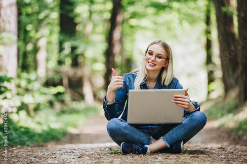 Young cheerful woman using laptop while sitting in forest