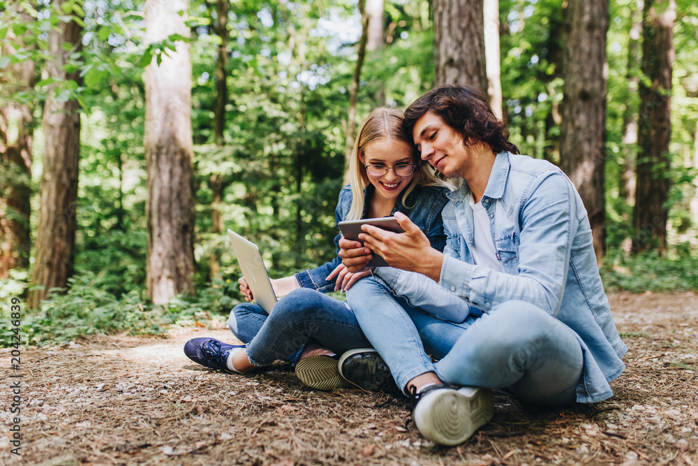 Young couple using laptop and tablet while sitting in forest Young couple using laptop and tablet while sitting in forest