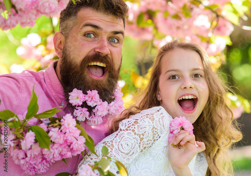Girl with dad near sakura flowers on spring day. Father and daughter on happy faces play with flowers and hugs, sakura background. Child and man with tender pink flowers in beard. Spring mood concept.