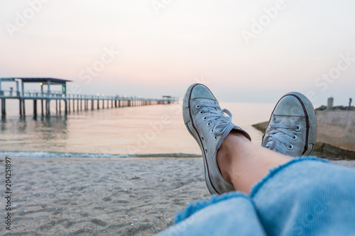 Legs of a woman in blue jeans and white shoes laying on beach beside the sea.
